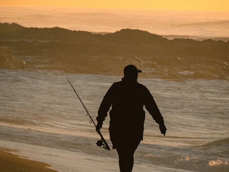 Silhouette of a fisherman carrying his rod along the shoreline at sunset