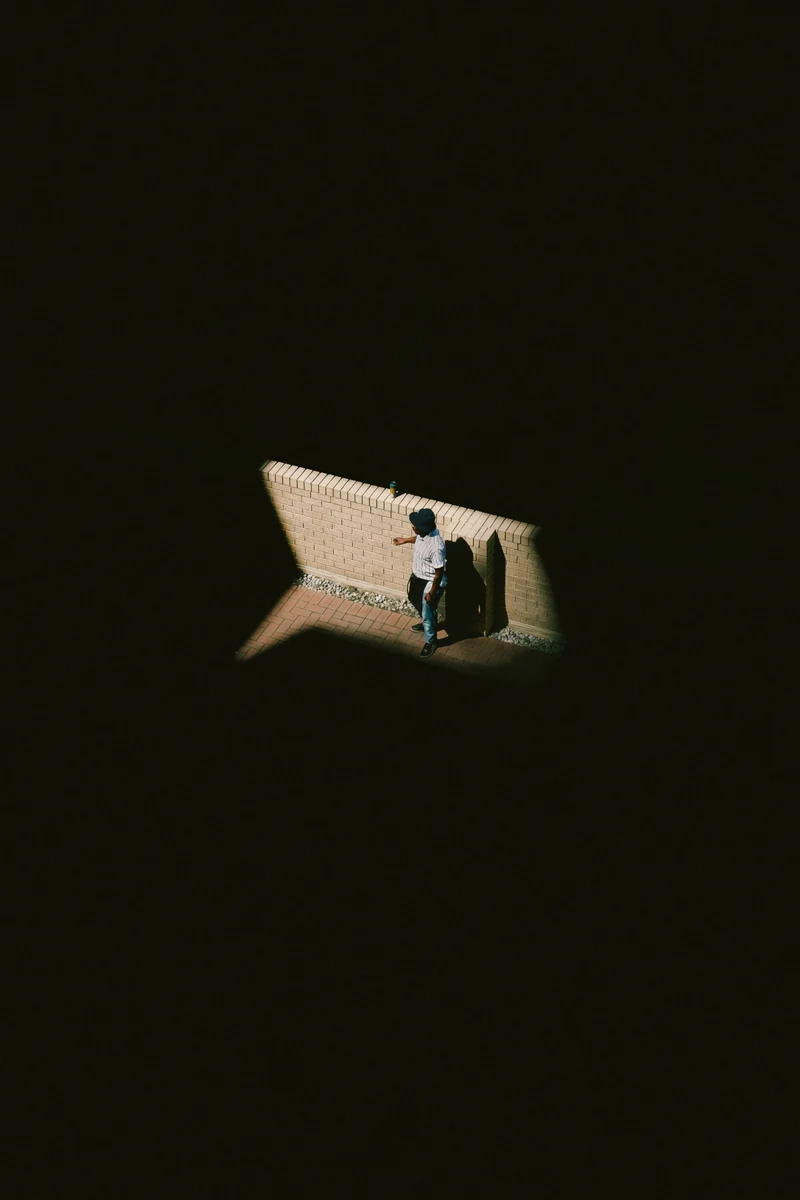 A man in a bucket hat standing in a patch of sunlight by a brick wall, seen from above