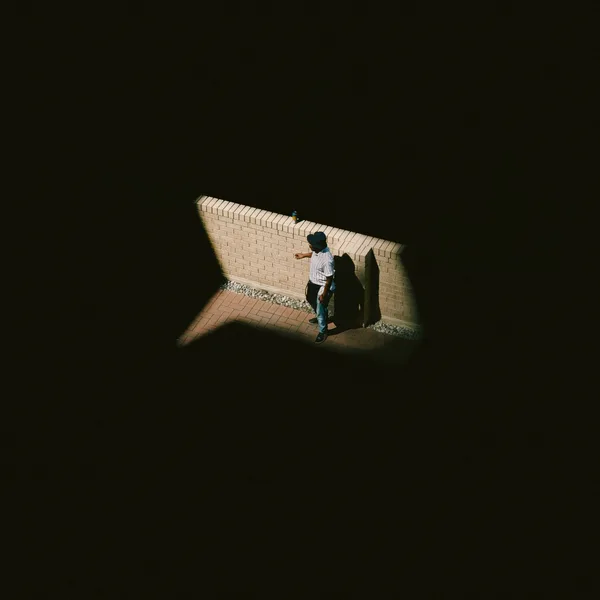 A man in a bucket hat standing in a patch of sunlight by a brick wall, seen from above