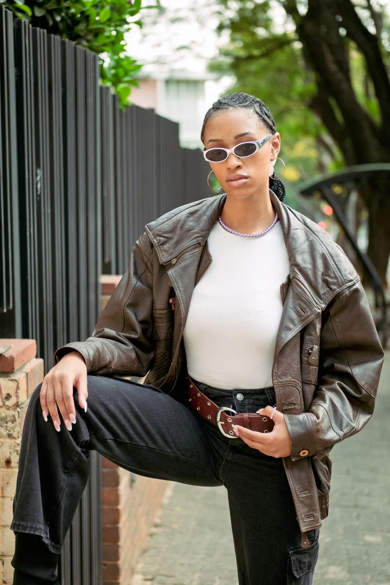 A woman in a brown leather jacket and white sunglasses posing by a black railing