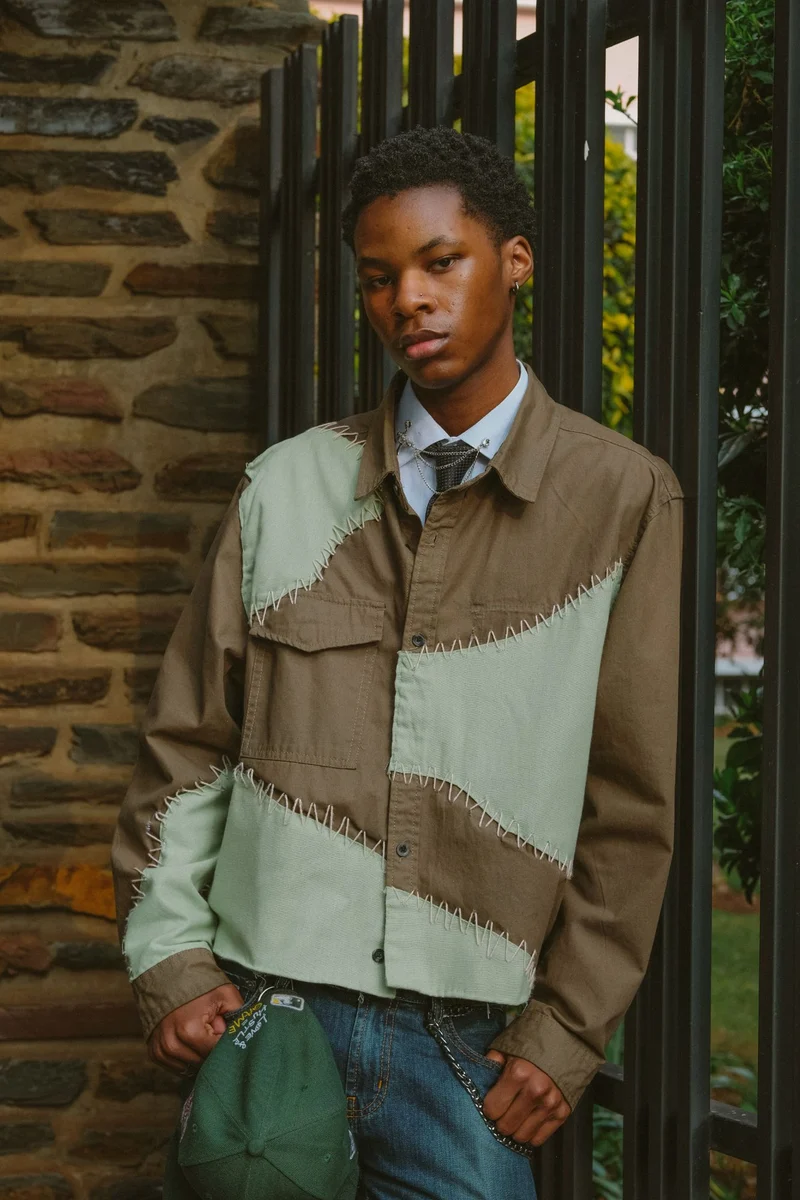 A young man in a patchwork shirt and tie leaning against a railing beside a stone wall