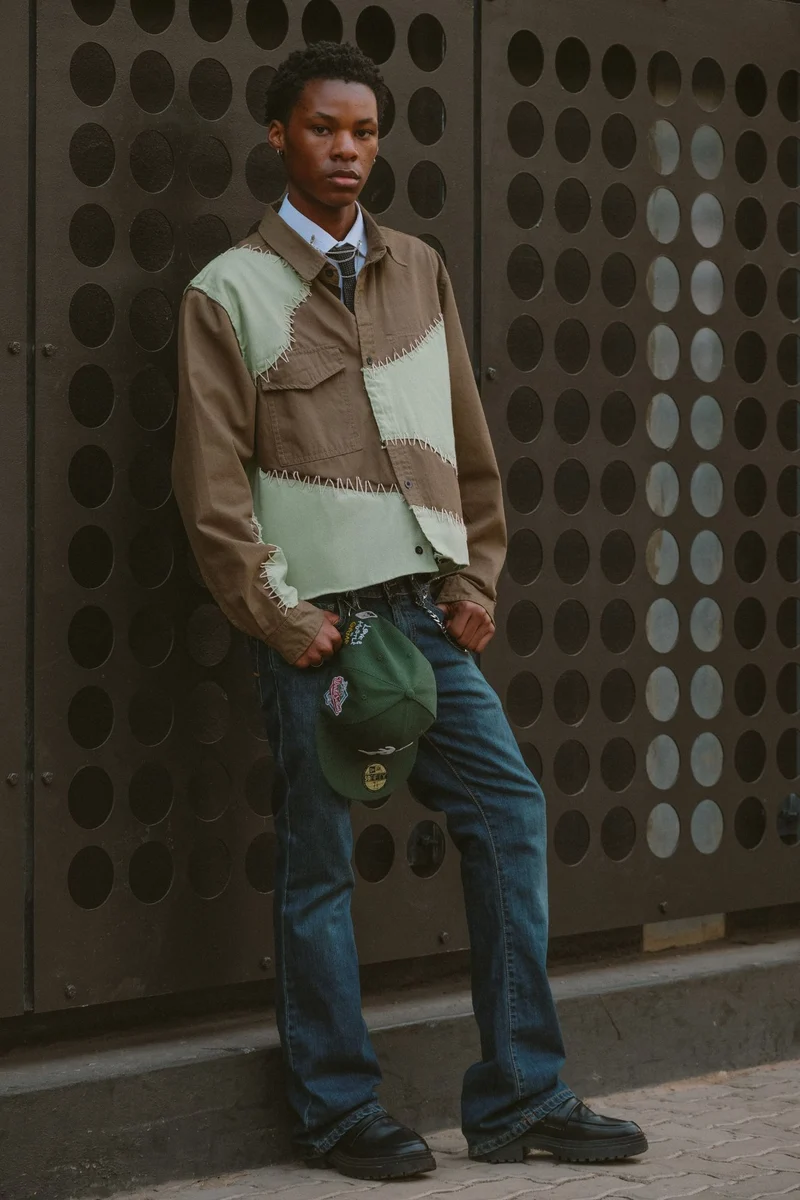 Full-length portrait of a young man in a patchwork shirt against a perforated metal wall