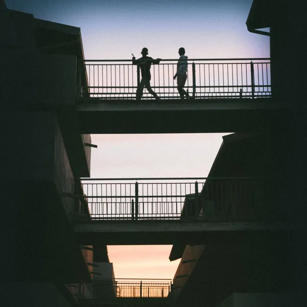 Two figures silhouetted on stacked walkways against a dusk sky
