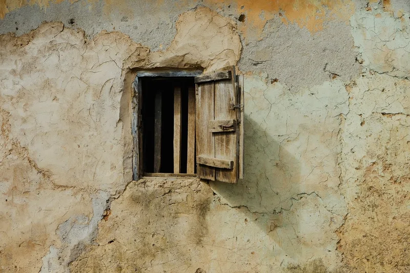 Small open window with a wooden shutter casting a long shadow on cracked plaster