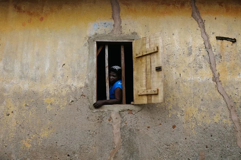 A woman in a blue top looking out of a barred window in a yellow wall