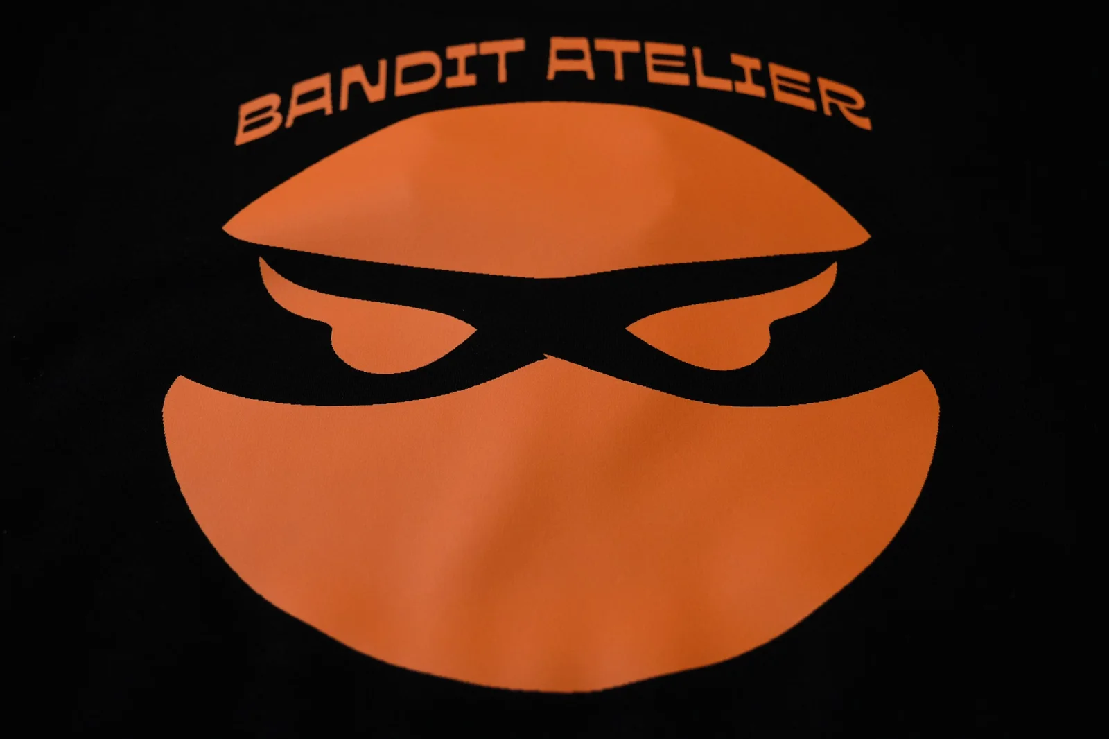 Bandit Atelier masked-face logo printed in orange on black fabric